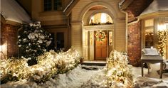 a house covered in snow with christmas lights on the front door and trees around it