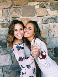 two beautiful women hugging each other in front of a brick wall with flowers on it