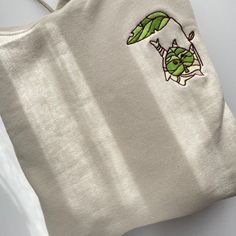 A beige Korok Hoodie by petimint, featuring an embroidered design of a small green leaf character hanging from a larger green leaf, is ideal for gaming enthusiasts. Positioned on a white surface, the stylish hoodie is bathed in natural light that casts subtle shadows across it. Winter Cotton Hoodie With Embroidered Patch, Cotton Hoodie With Embroidered Patch For Streetwear, Casual Hoodie With Embroidered Patch For Winter, Casual Winter Hoodie With Embroidered Patch, Green Cotton Hoodie With Embroidered Graphics, Hooded Cotton Tops With Embroidered Graphics, Casual Hoodie With Embroidered Patch, Green Casual Hoodie With Custom Embroidery, Casual Green Hoodie With Custom Embroidery