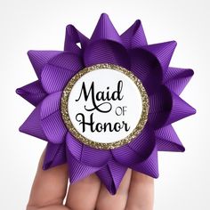 a hand holding a purple ribbon with the words maid of honor written on it in black ink