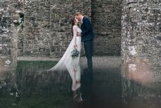 a bride and groom are kissing in front of an old stone wall with water reflecting them