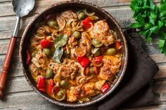 a skillet filled with chicken and vegetables on top of a wooden table next to a spoon