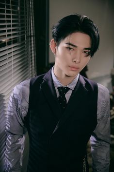 a young man wearing a black vest and tie in front of a window with blinds