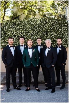 a group of men standing next to each other wearing suits and bow ties in front of a hedge