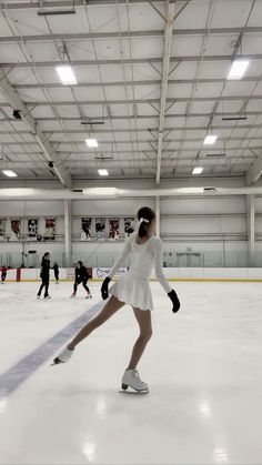 a woman skating on an ice rink in a white dress and headphones, with other people behind her