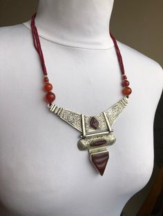 A beautiful and unique bohemian necklace in an eastern style, with deep carnelian and seed beads. This beautiful piece is closed with a toggle clasp. Three strands of black matte seed beads lead from it to the main piece - an angular metal plate, holding aother unusually shaped element - an oval merged with a triangle. There are three carnelian gemstones sitting down the middle of the necklace, framed by the curls and twigs of the metal embossing. This beautiful, elegant piece will look magnific Bohemian Silver Beaded Necklaces With Carnelian, Tuareg Jewelry, Metal Embossing, Carnelian Necklace, Coral Bracelet, Bohemian Necklace, Boho Gifts, Metal Plate, Bohemian Jewelry