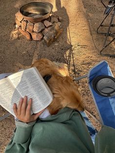 a person reading a book with a dog laying on their back next to them and a fire pit in the background