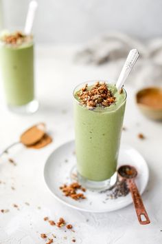 two glasses filled with green smoothie sitting on top of a white plate next to some spoons