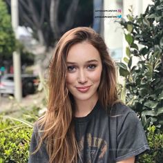 a woman with long hair and blue eyes posing for a photo in front of bushes