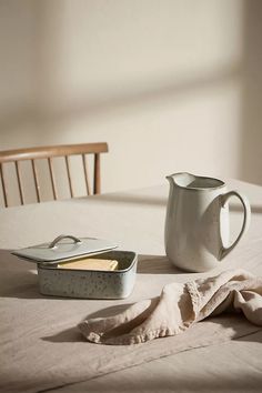 a table topped with a white pitcher next to a container filled with food on top of a bed