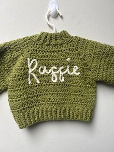 a green knitted sweater with the word roggie on it hanging from a hook