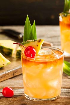 two glasses filled with pineapple and cherries on top of a wooden table next to sliced pineapples