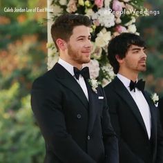 two men in tuxedos standing next to each other with flowers on the wall behind them