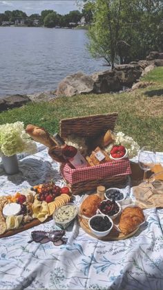 a picnic with bread, fruit and cheeses on a blanket next to the water