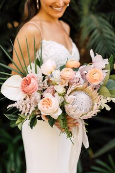 a bride holding a bouquet of flowers in her hands and smiling at the camera with palm trees behind her