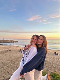 two young women are hugging on the beach at sunset, with people in the background