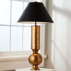 a gold lamp with a black shade on it in front of a white wall and window