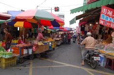 people are shopping at an outdoor market with umbrellas and fruit stands in the background