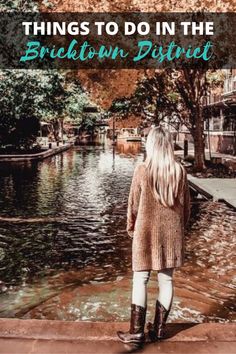 a woman standing on the edge of a pond looking at trees and buildings in autumn