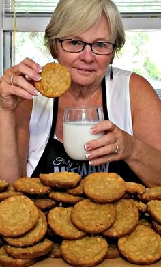 a woman holding a cookie next to a pile of cookies and a glass of milk