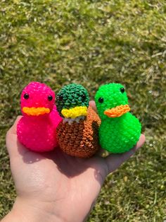 three small crocheted ducks sitting on top of a green grass covered field next to a person's hand