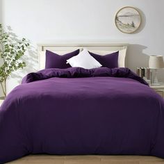 a bed with purple comforter and pillows in a white room next to a potted plant