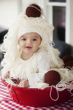 a baby wearing a white hat and dress sitting in a red bucket on a checkered table cloth