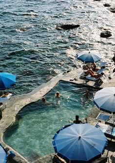 people are swimming in an outdoor pool next to the ocean with umbrellas and chairs