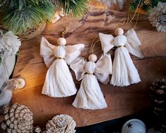 three white angel ornaments on a wooden table with pine cones and evergreen branches in the background