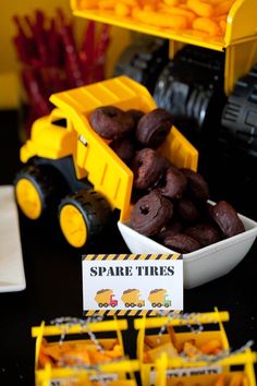 a table topped with yellow toy trucks next to orange candies and cupcakes