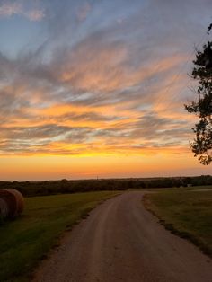 a dirt road with hay bales in the foreground and a sunset behind it