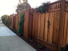 a wooden fence next to a sidewalk with plants growing on the top and bottom of it