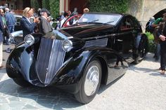 an old black car parked on the side of a road with people standing around it