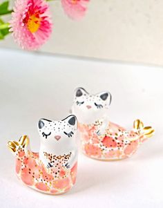 two white and pink cat figurines sitting on top of a table next to flowers