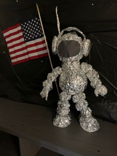 an astronaut doll made out of tin foil with the american flag in the back ground