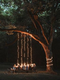 a group of people sitting around a table under a tree with lights strung from it