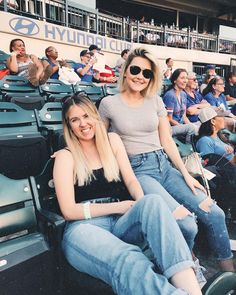 two women are sitting in the bleachers at a baseball game, one is wearing sunglasses