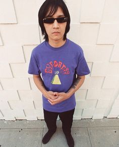 a man with sunglasses on standing in front of a white wall wearing a purple shirt and black pants