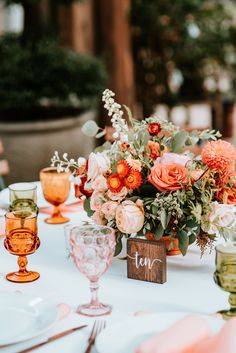 an arrangement of flowers and greenery on a white table cloth with place settings for dinner