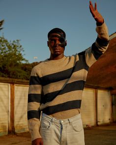 a man in striped sweater waving at the camera with his hand up and wearing white jeans