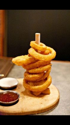 a stack of onion rings sitting on top of a wooden board next to dipping sauces