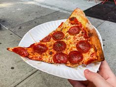 a slice of pepperoni pizza on a paper plate with someone's hand holding it