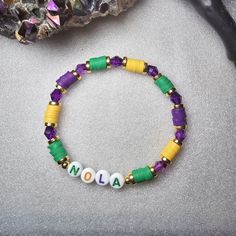 The New Orleans NOLA Mardi Gras Beaded Word Bracelet was made using 6mm crystal bicone beads, circular spacer beads, and heishi clay beads. The letters "NOLA" are spelled out on white round letter beads in purple, yellow, and green writing.  Bracelets are all HANDMADE with love and care by myself, Candice Marie, and they are always limited edition and one-of-a-kind. ❤️ The bracelet is stretchy and stackable and looks great stacked with other Candice Marie Bead Co. beaded word bracelets in my sho Adjustable Beaded Spiritual Name Bracelet, Spiritual Beaded Adjustable Name Bracelet, Stretch Bracelet With 8mm Heishi Beads, Adjustable Beaded Bangle Bracelets With Letter Beads, Spiritual Beaded Name Bracelet With Round Beads, Purple Beaded Heishi Beads Bracelets, Spiritual Beaded Name Bracelet, Beaded Name Bracelet With Round Beads For Friendship, Adjustable Hand-strung Name Bracelet With Round Beads