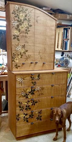 a dog standing next to a wooden cabinet with flowers painted on the doors and drawers