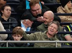 two men are sitting in the stands at a tennis game and one man is holding up his cell phone