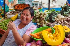 a woman smiles at the camera while surrounded by vegetables and fruit for sale in a market