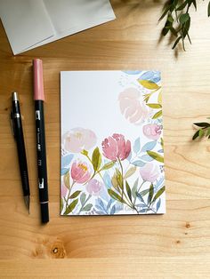 a notepad with watercolor flowers on it next to a pen and flower arrangement