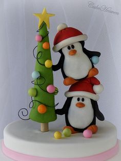 two penguins standing next to each other near a small christmas tree on top of a white cake