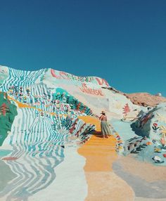 a woman standing in the middle of a desert with painted walls and mountains behind her