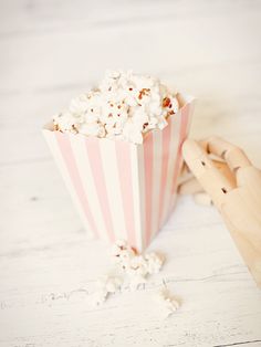 a pink and white striped popcorn bucket with wooden spoons next to it on a white table